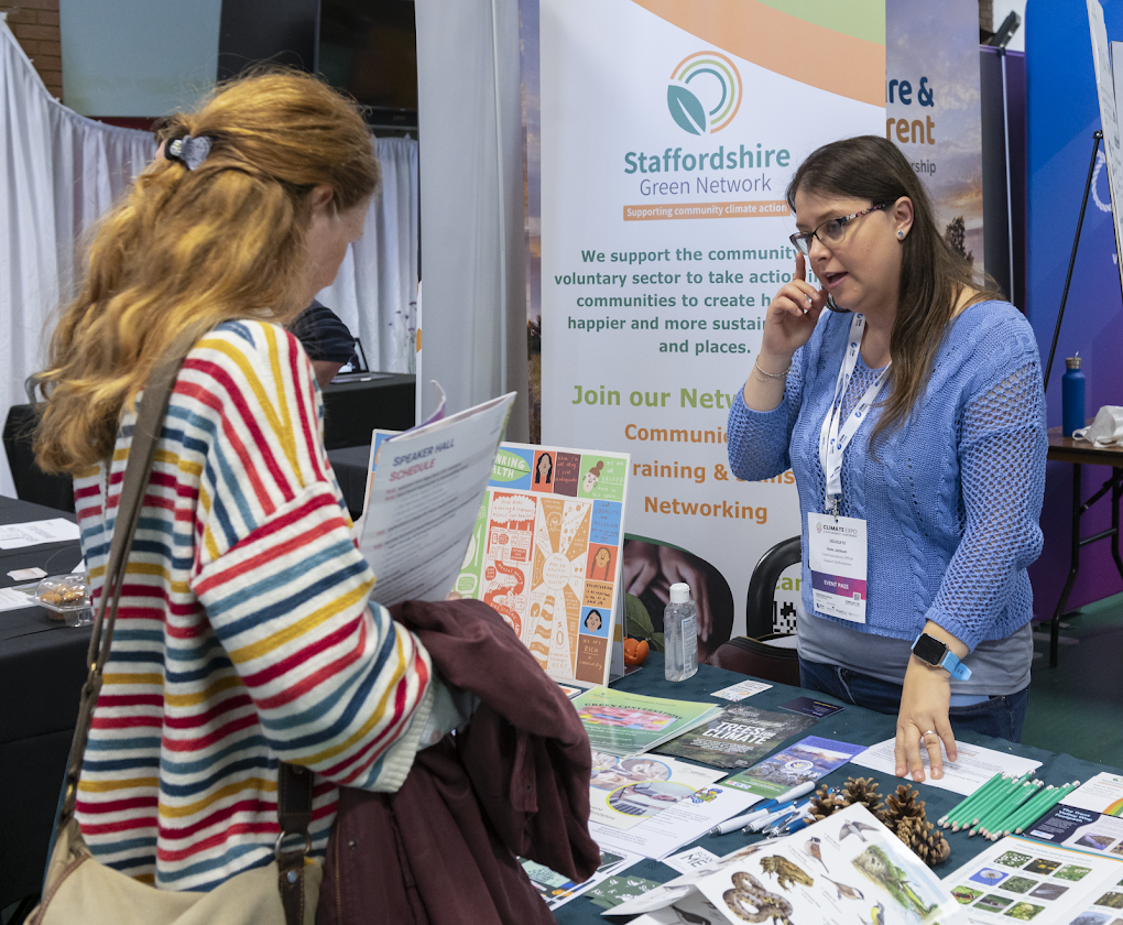 Two women stand at a stall full of colourful leaflets, with a banner saying 'Staffordshire Green Network' in the background