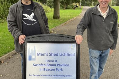 Two men standing behind an advertising board in a park. The board gives the contact details for Men's Shed Lichfield