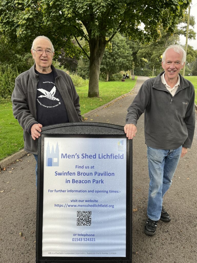 Two men standing behind an advertising board in a park. The board gives the contact details for Men's Shed Lichfield